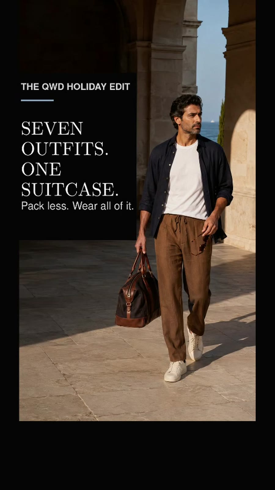 A man wearing a navy layer, white T-shirt, tobacco trousers and white sneakers on holiday