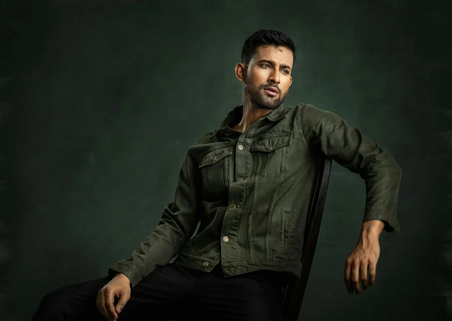 A seated man wearing an olive trucker jacket and dark trousers against a green background