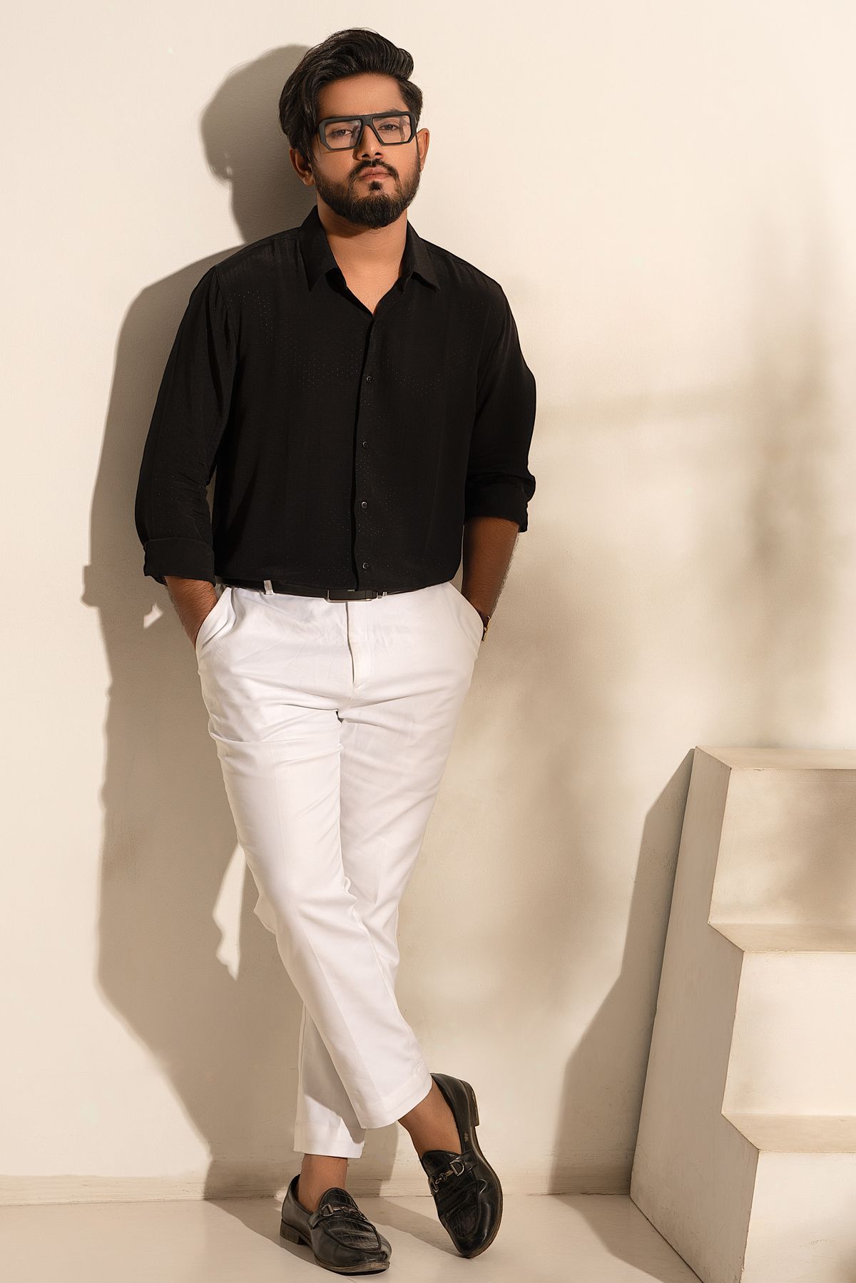 A man wearing a black shirt, cropped white trousers and black bit loafers against a cream studio wall